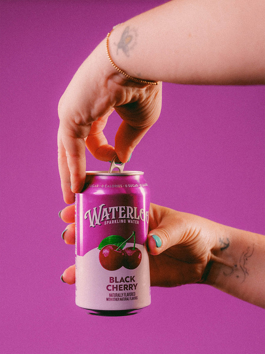 Close-up of hands opening a can of Waterloo Black Cherry sparkling water against a vibrant purple background.