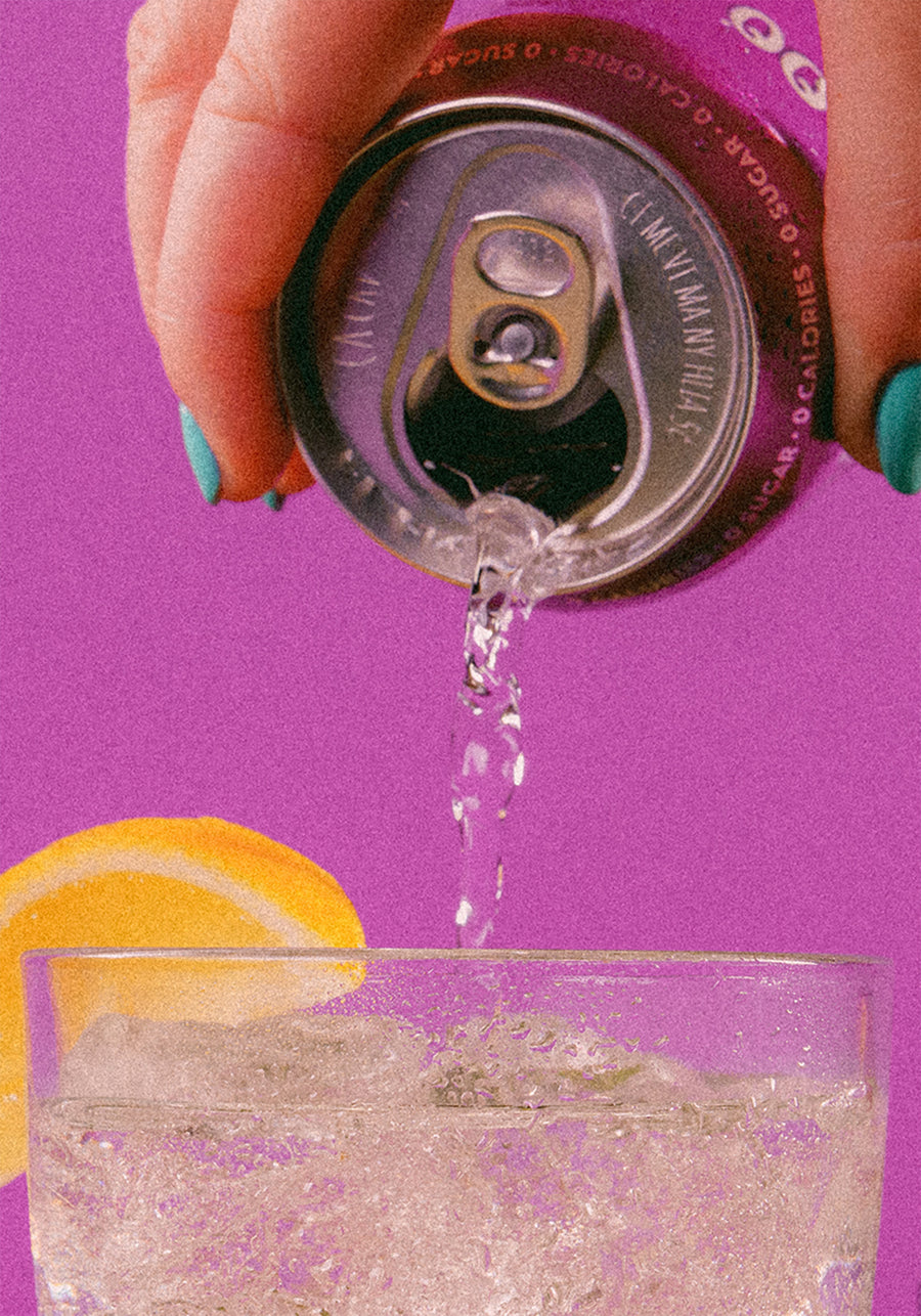 Hand pouring a sparkling drink from a can into a glass, with a lemon slice in the background on a pink surface.