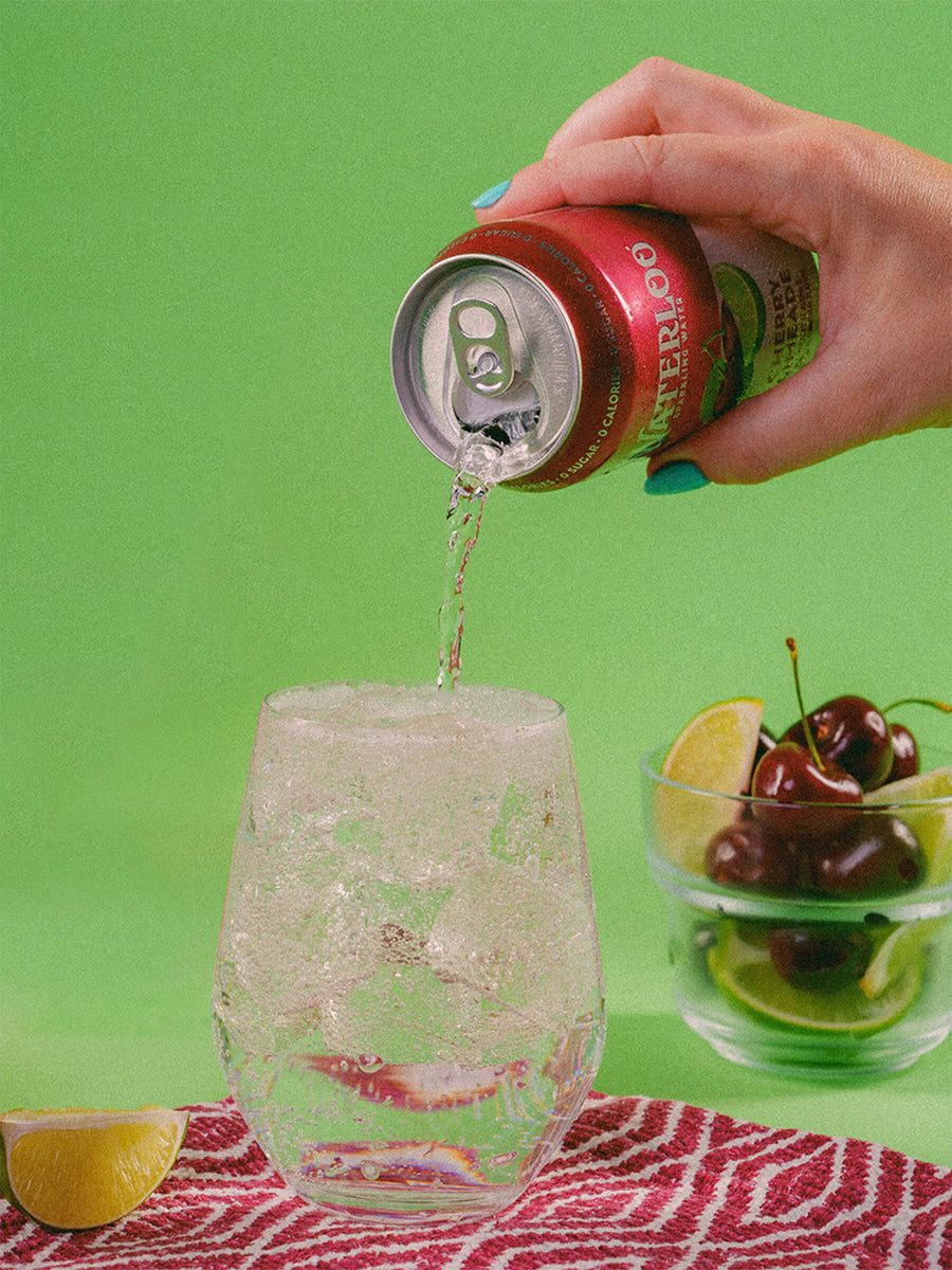 A hand pouring a sparkling drink from a can into a glass, garnished with lime and cherries on a vibrant green background.
