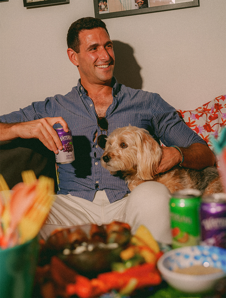 Man smiling while holding a drink, enjoying time with a dog on the couch amid colorful snacks and decor.