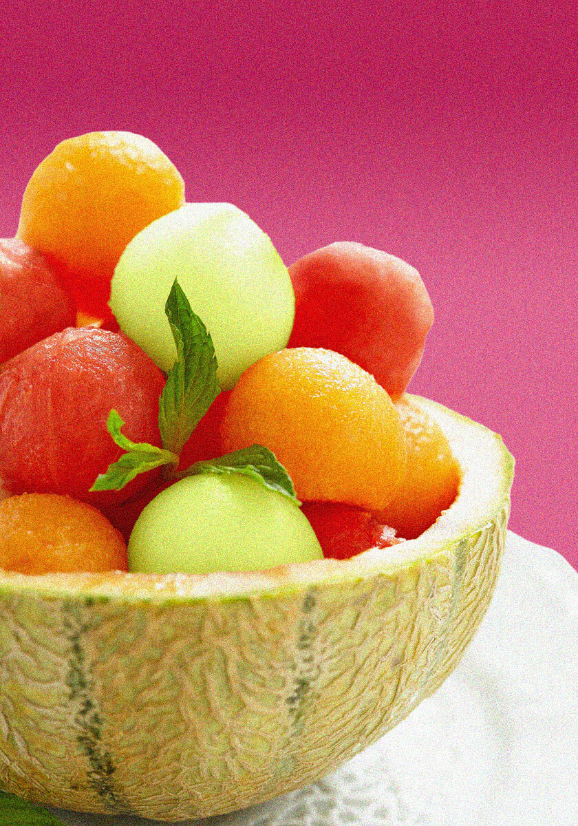 Colorful fruit salad of melon balls served in a half cantaloupe, garnished with mint leaves on a pink background.