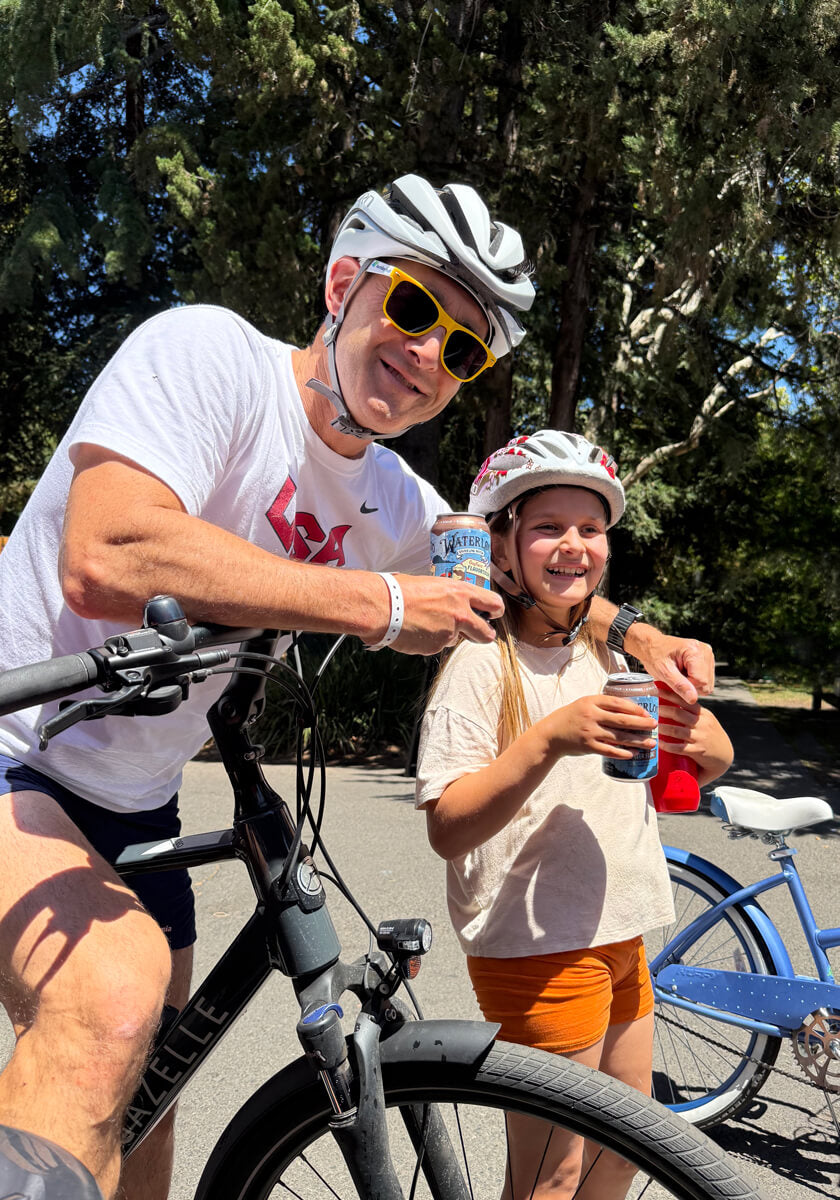 A father and daughter enjoying a sunny day biking, both holding drinks and smiling, showcasing family bonding.