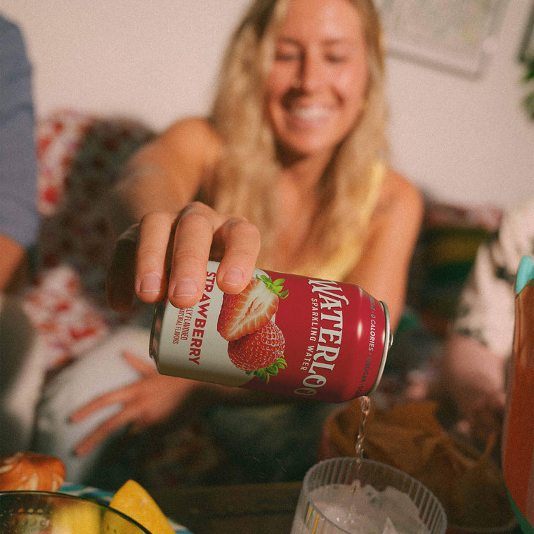 A woman pouring Strawberry Waterloo sparkling water into a glass filled with ice, smiling in a cozy setting.
