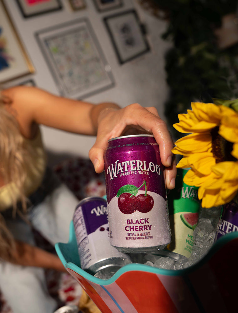 A hand holding a can of Waterloo Black Cherry sparkling water beside floral decor and ice-filled bucket.