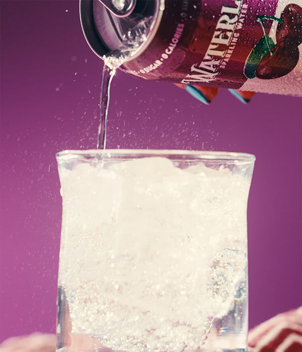 A can of sparkling water pouring into a glass filled with ice, against a purple background.