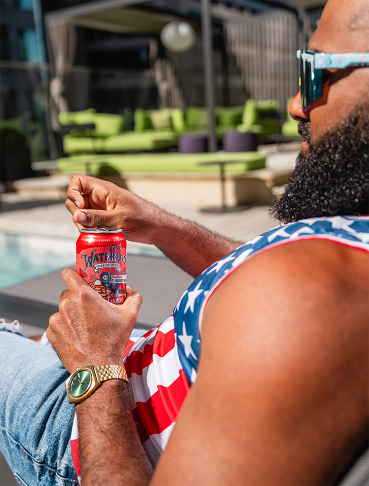 Man in patriotic tank top enjoying a can of Watermelon sparkling water by the poolside on a sunny day.