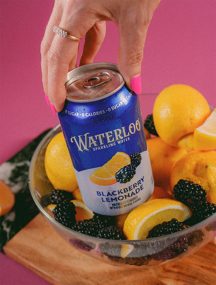 A hand holding a can of Waterloo Blackberry Lemonade sparkling water, with lemons and blackberries in a bowl.