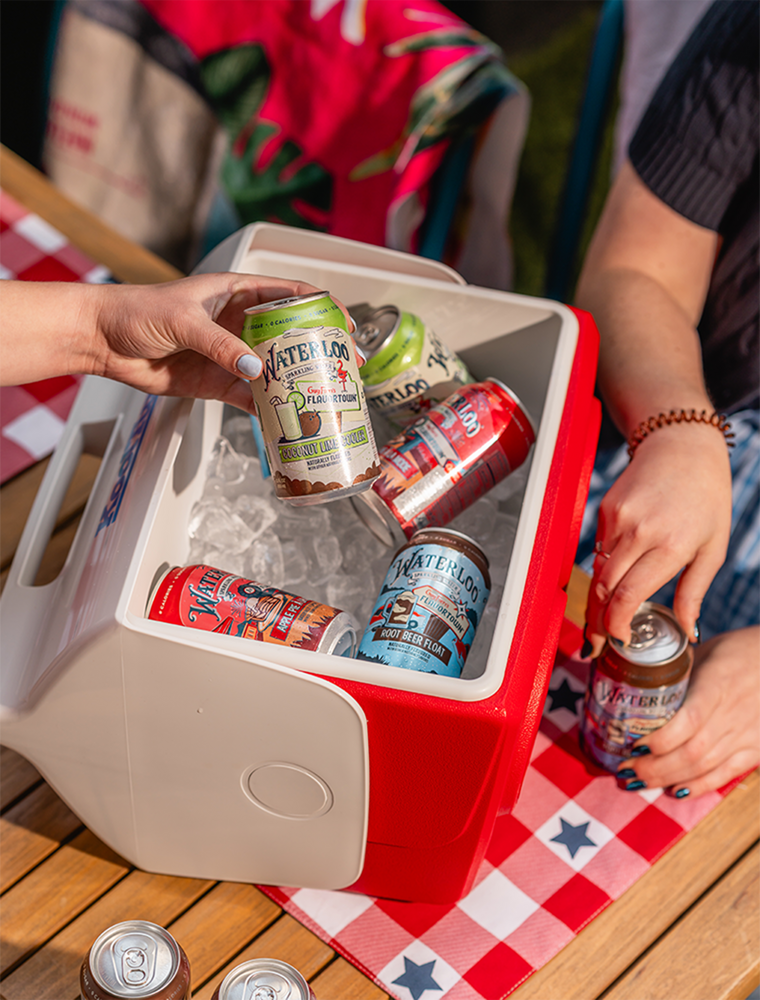 Hand picking up a can of Waterloo sparkling water from an ice-filled cooler at a picnic table.
