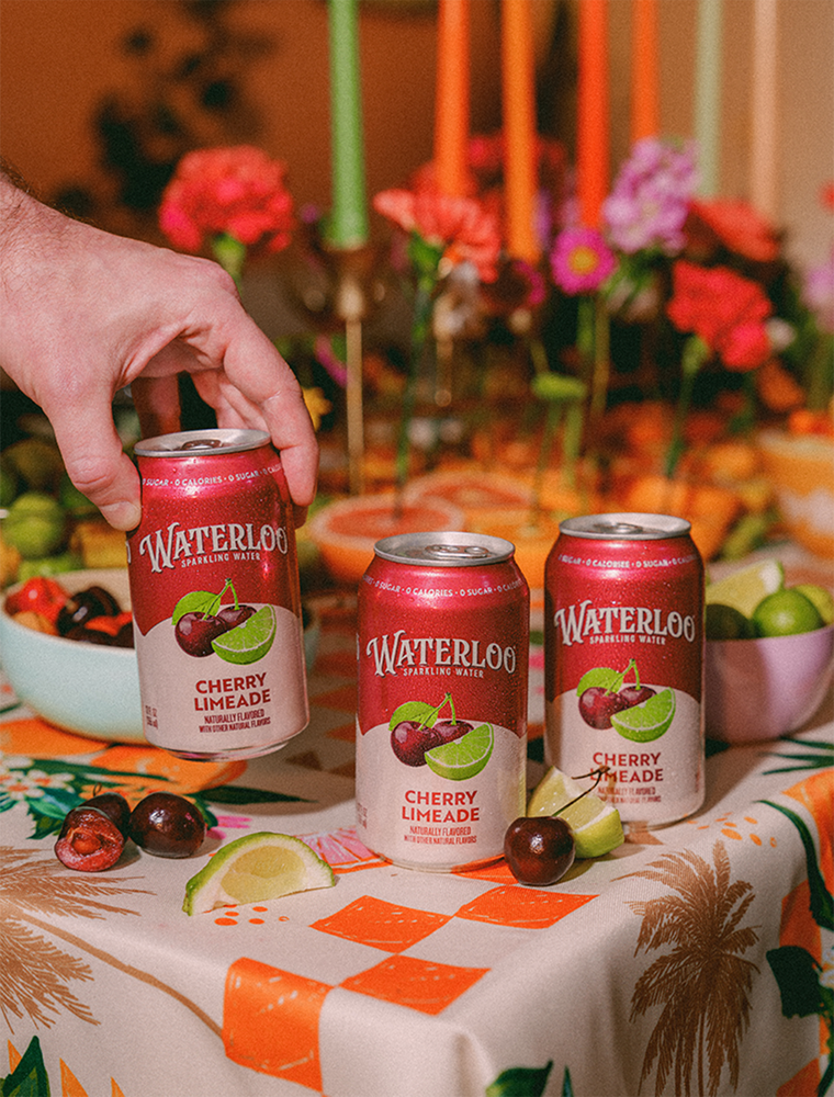 A hand reaching for a can of Waterloo Cherry Limeade on a colorful table with festive decorations.