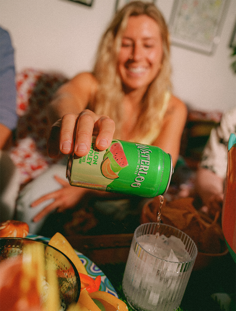 A woman joyfully pours watermelon-flavored sparkling water into a glass filled with ice.