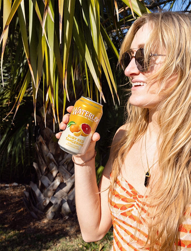 Woman enjoying Waterlog Ruby Red Tangerine sparkling water in a sunny outdoor setting with palm trees.