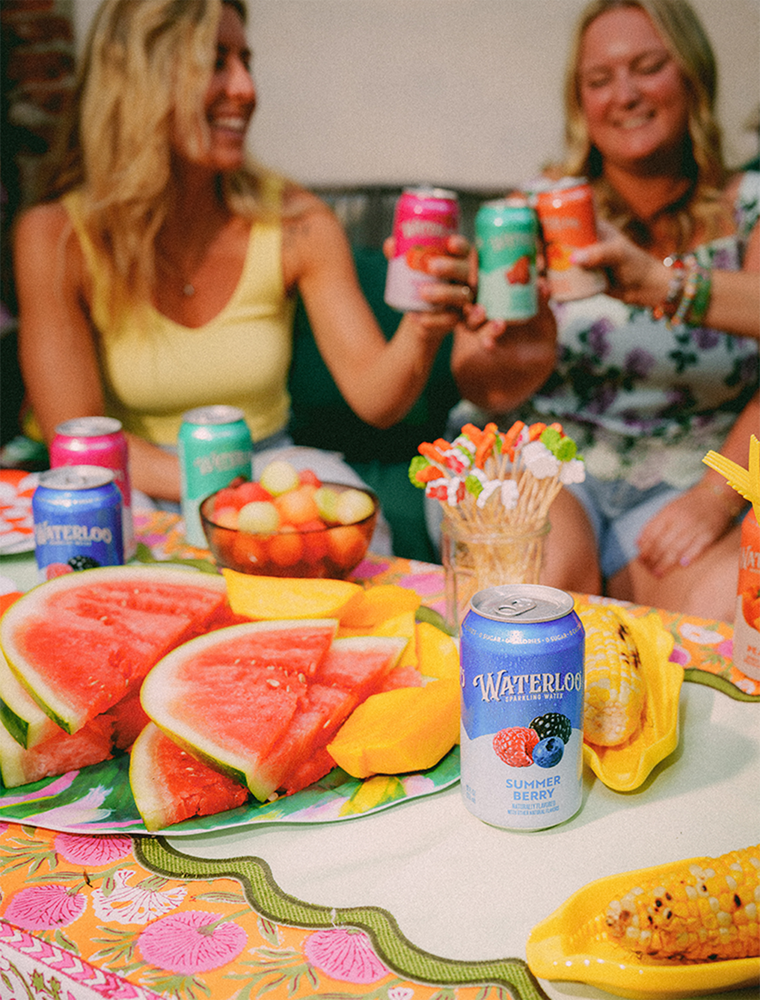 Two women toasting with colorful drinks at a summer gathering, featuring fresh watermelon and snacks on the table.