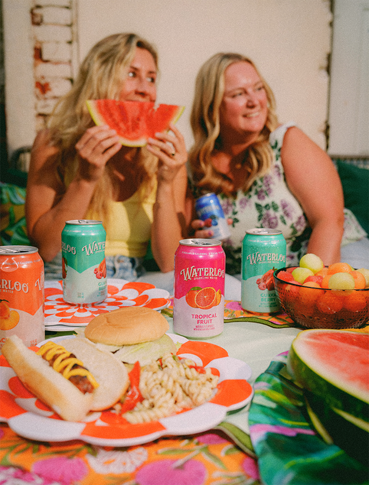 Two women enjoying a summer picnic with watermelon, snacks, and colorful Waterloo beverages on a vibrant table.