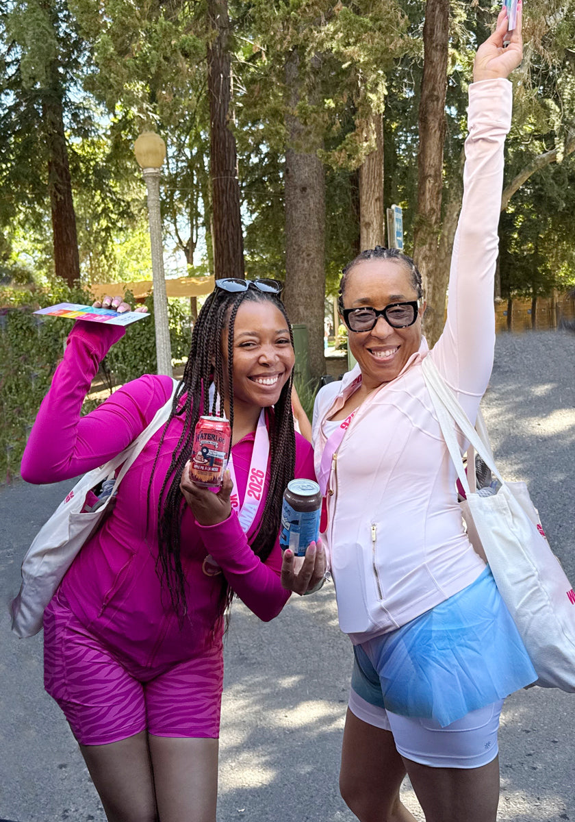 Two smiling women in pink athletic wear holding drinks outdoors, celebrating in a park setting.