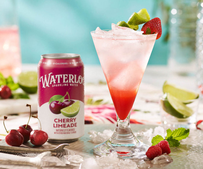 A vibrant glass of cherry limeade cocktail with ice, garnished with strawberries and lime next to a Waterloo can.
