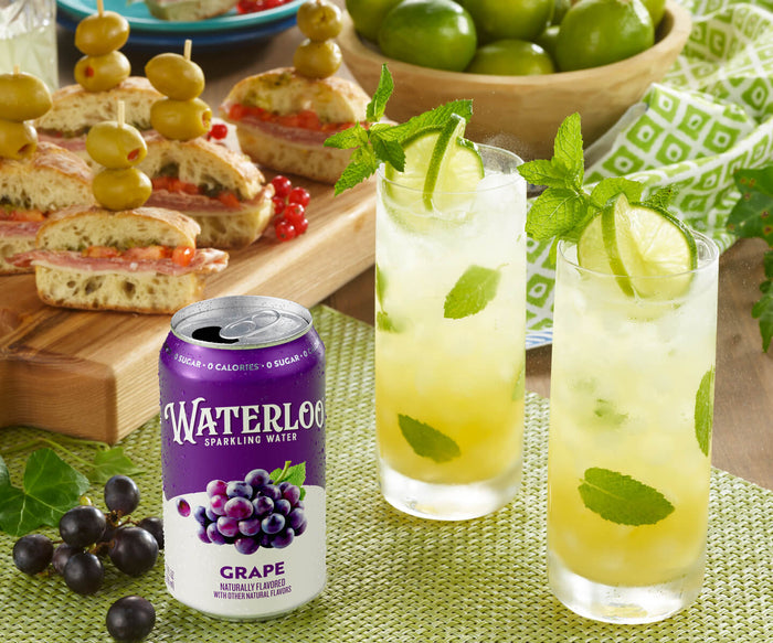Refreshing drinks with mint and lime beside Waterloo Grape sparkling water and gourmet sandwiches on a wooden platter.
