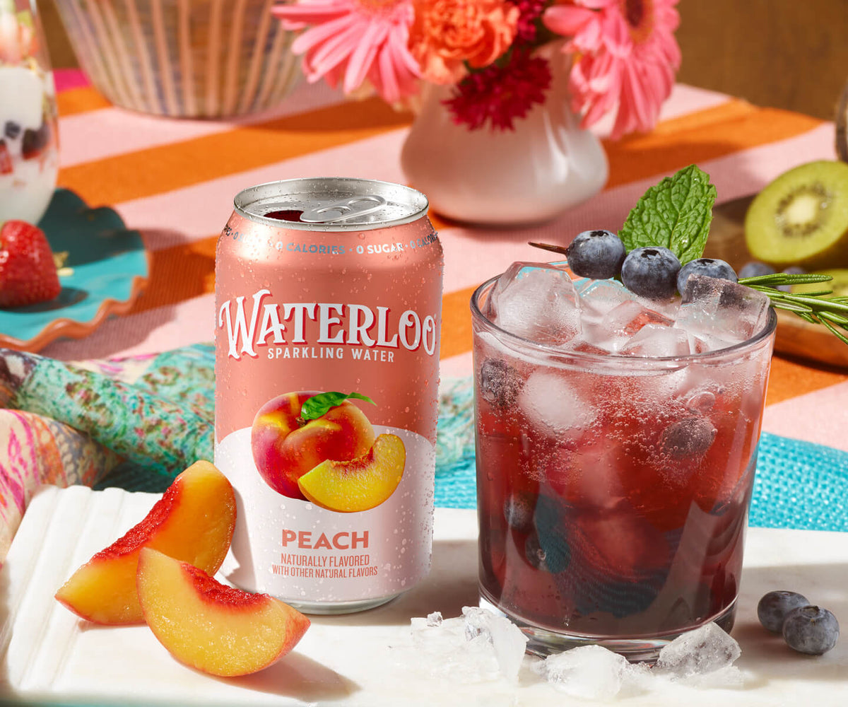 Waterloo Peach sparkling water can beside a glass filled with ice, blueberries, and mint on a colorful table setting.