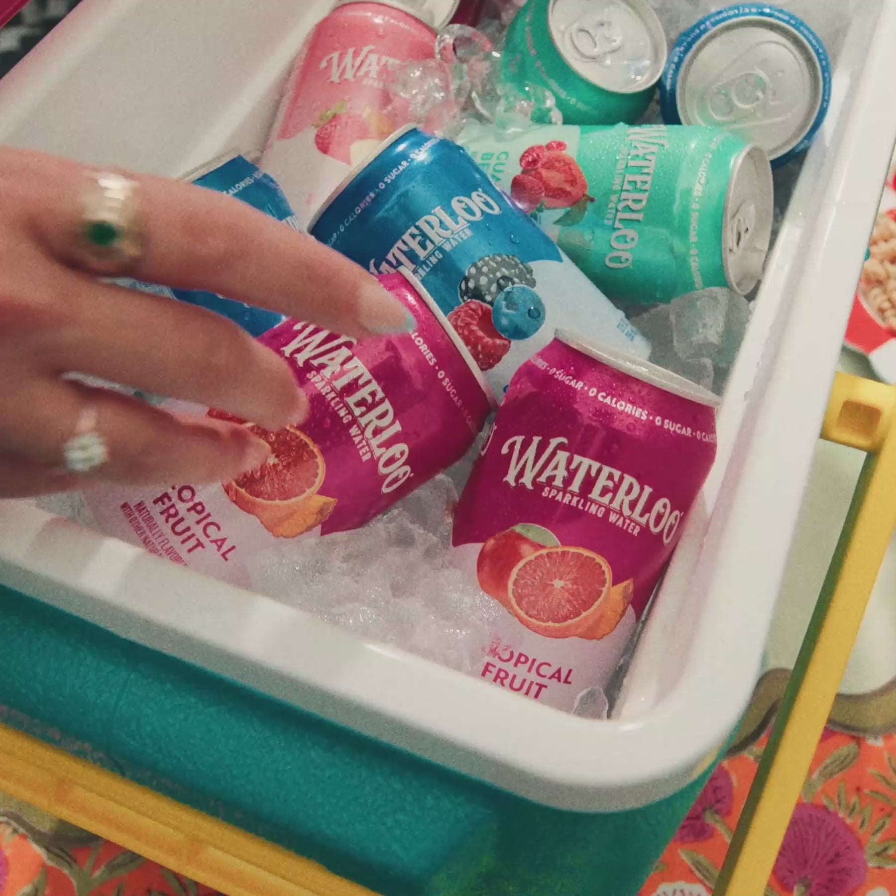 A hand reaching for colorful cans of Waterloo sparkling water, featuring Tropical Fruit flavor and ice.